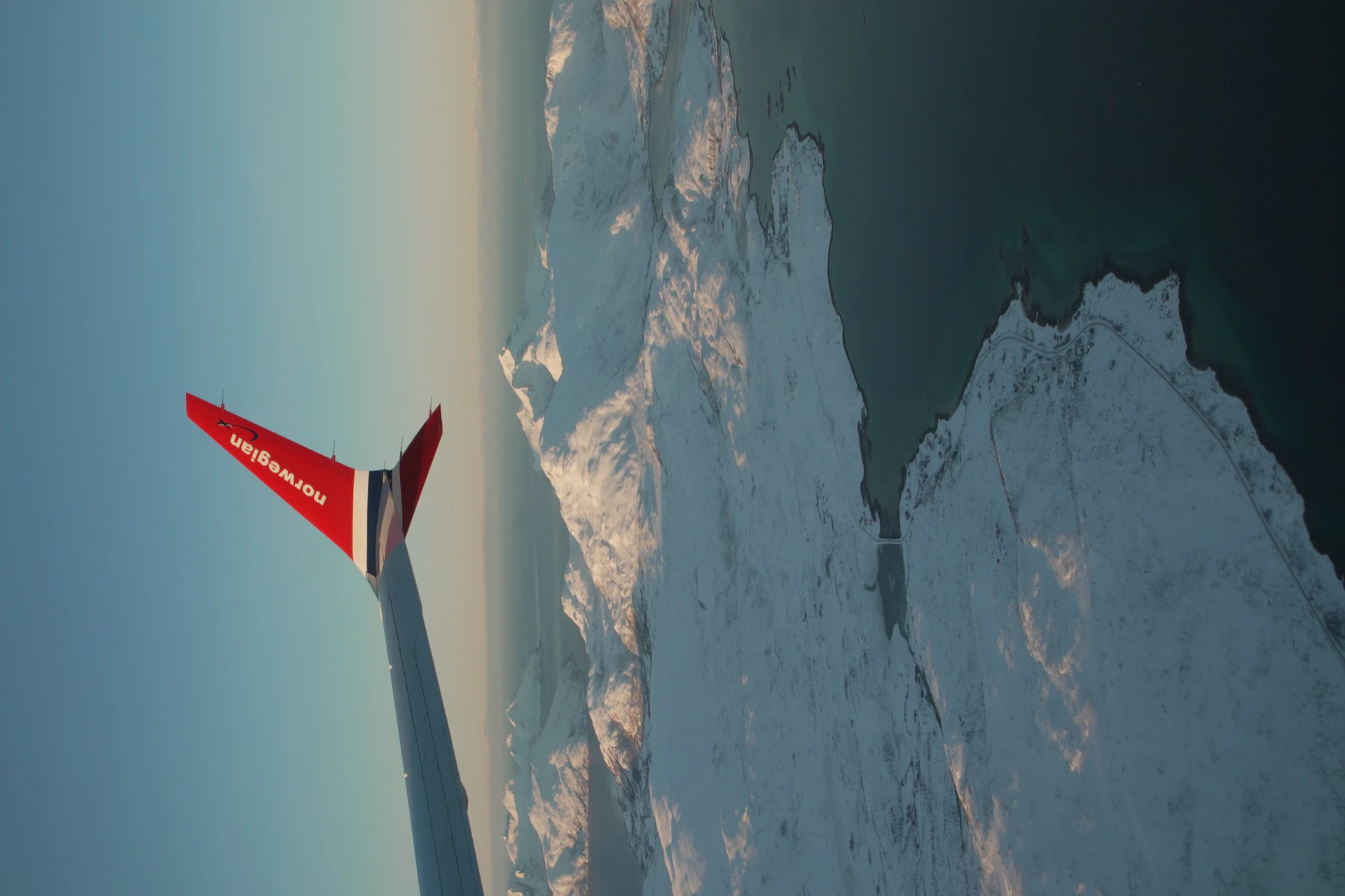 Looking down from a Norwegian Air flight at snow-capped mountains and dark water.